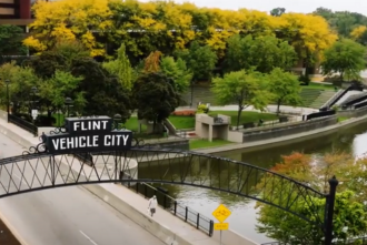 Overpass sign reading "FLINT VEHICLE CITY" with trees, river, walking path, and city buildings in the background
