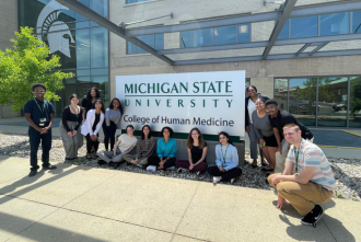 Group of college interns outside a large  building with glass windows and a sign that says, "Michigan State University College of Human Medicine"