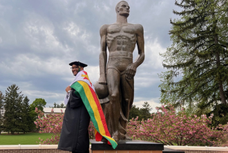 Male student wearing graduation gown posing with large bronze Spartan statue
