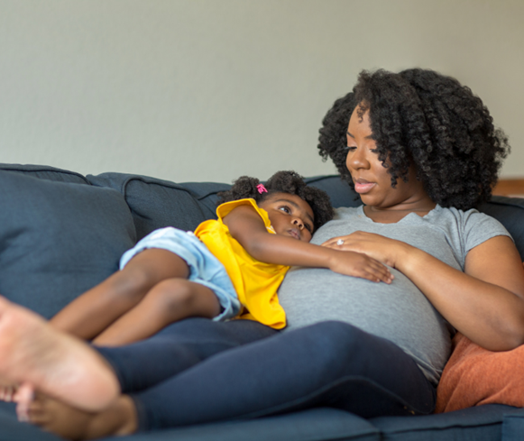 A pregnant woman and her young child lay next to each other on a couch. Both of them have their hand on the mom's stomach.