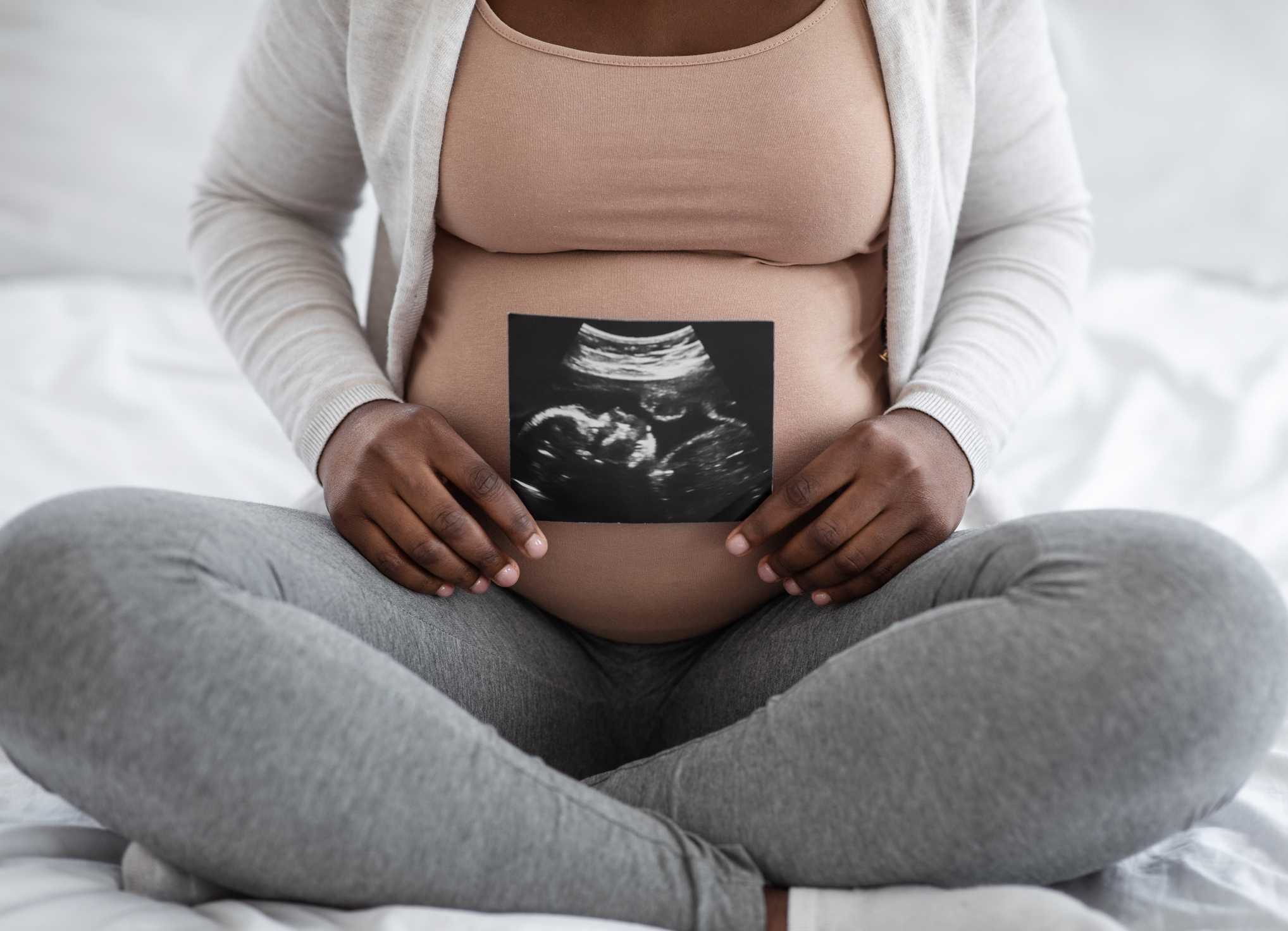 A pregnant woman visible from the neck down sitting cross-legged holding an ultrasound photo.
