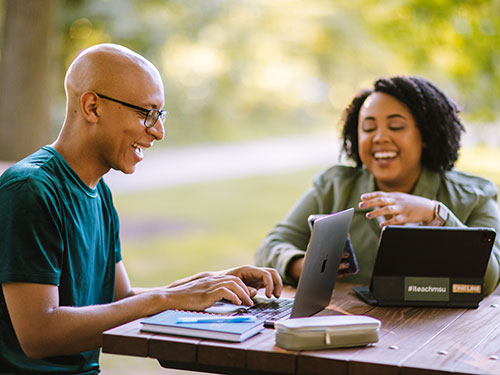 Two students sitting at a table outdoors, smiling and using a laptop and tablet