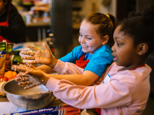 Two children are engaged in a cooking activity, with their hands in a mixing bowl, smiling and focused on the task