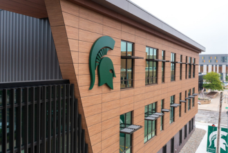 Exterior photo of the Charles Stewart Mott Department of Public Health building expansion focusing on the top left MSU Helmet icon hanging on the building