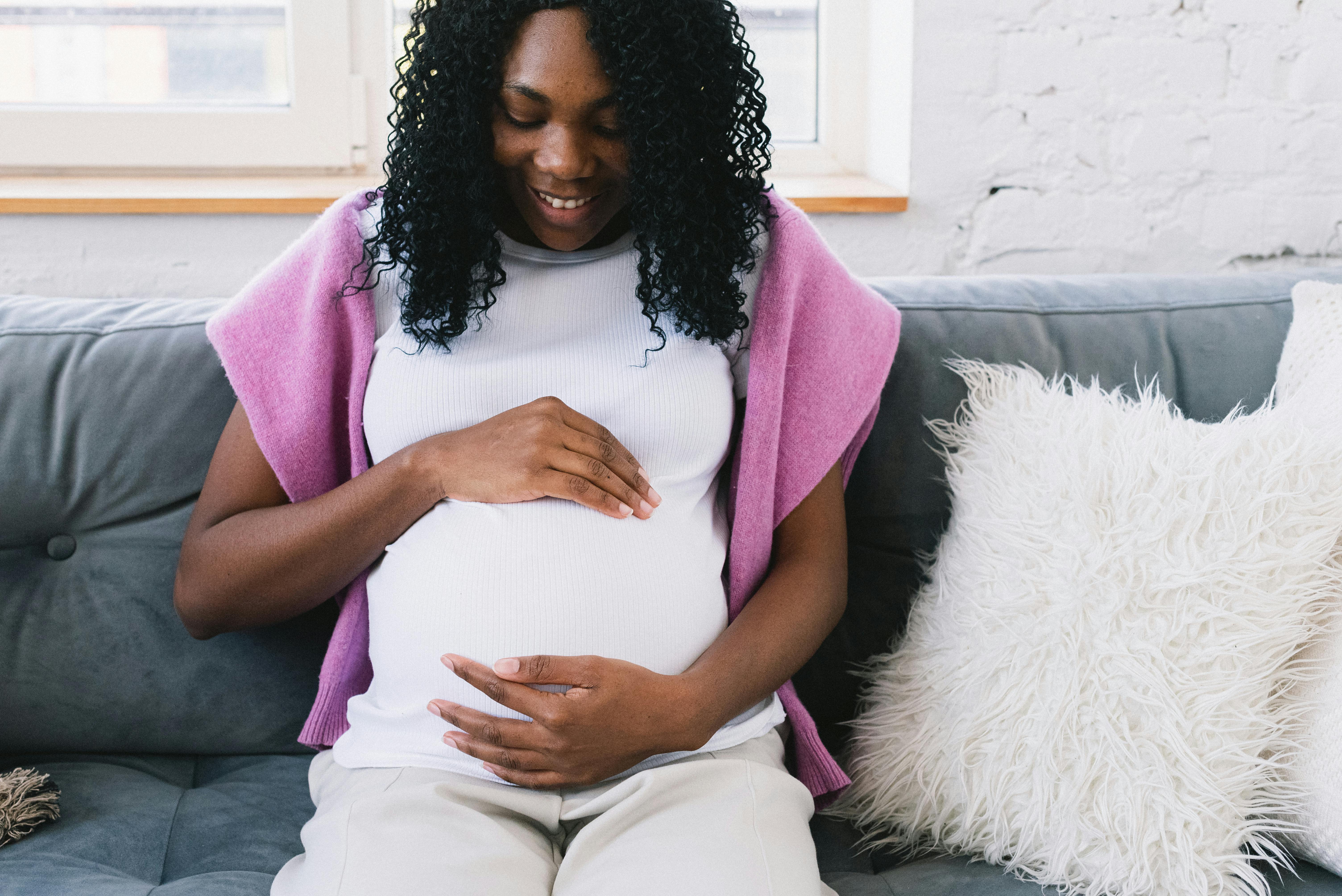 A pregnant woman sitting on a couch holding her stomach while looking down and smiling.